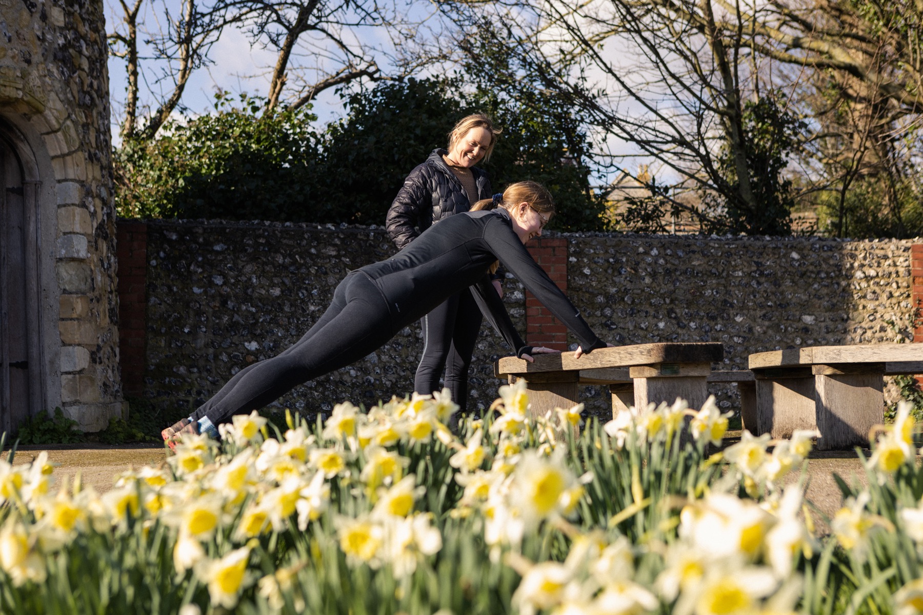Two women doing an incline exercise against a flint wall, a bank of daffodils in sharp focus in the foreground