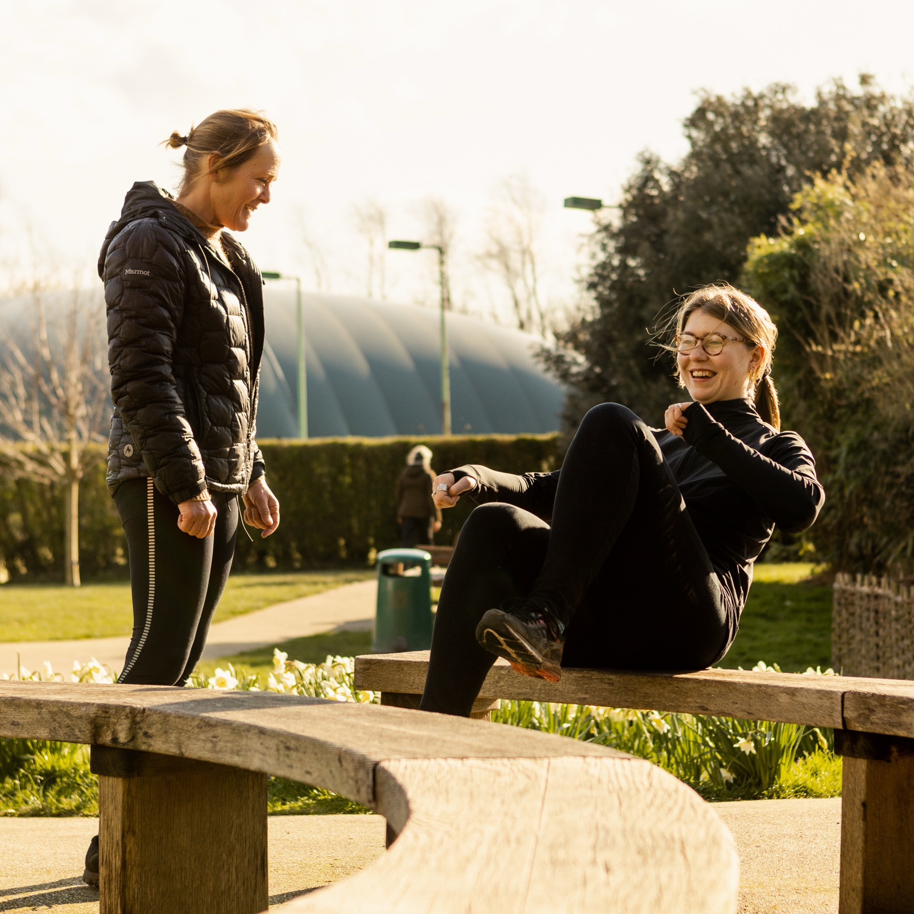 Two women on a curved wooden bench doing seated exercises, an open playing field and an inflated sports dome behind