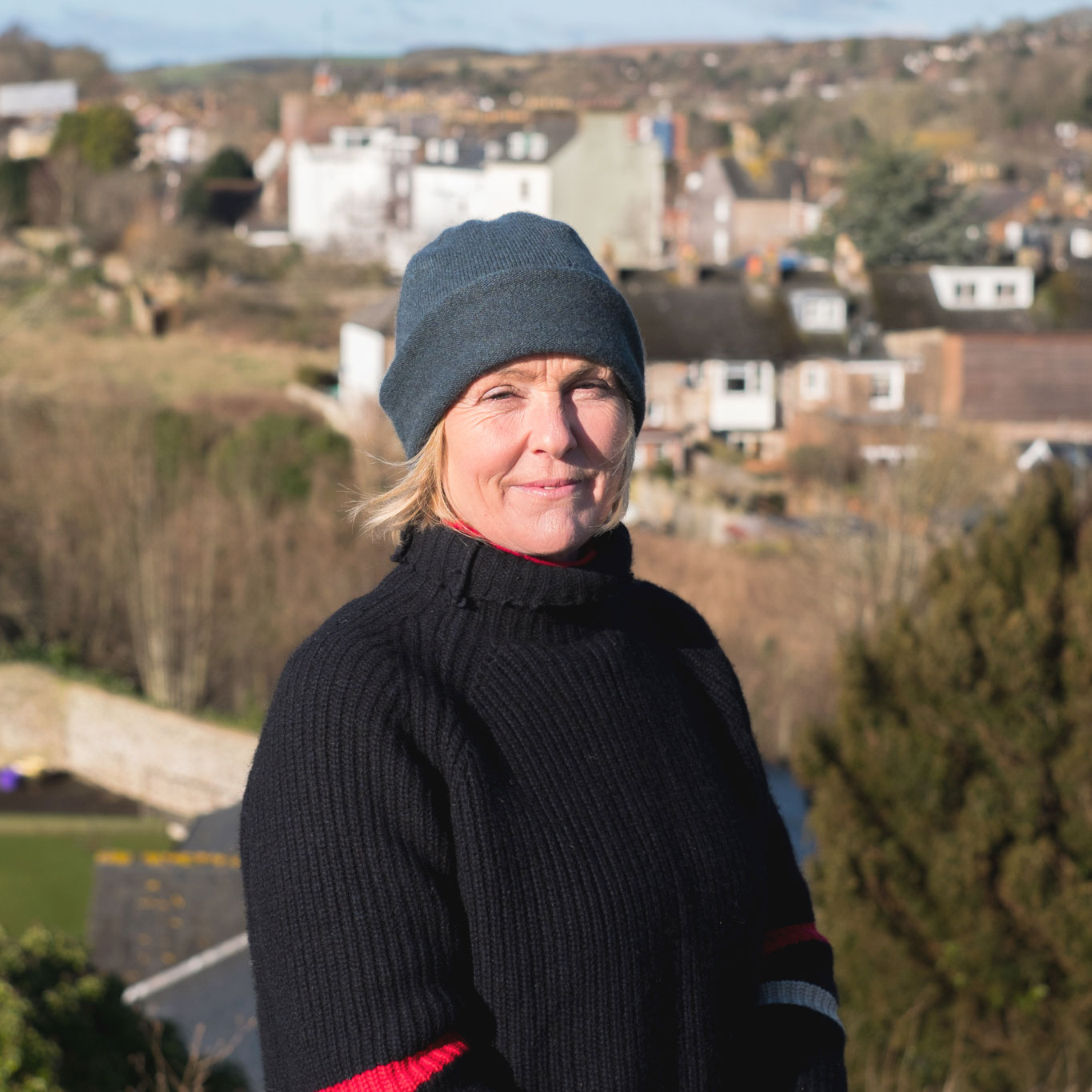 Maria Judd, head and shoulders, in a dark grey beanie and black roll-neck, the rooftops of Lewes and the Downs behind her