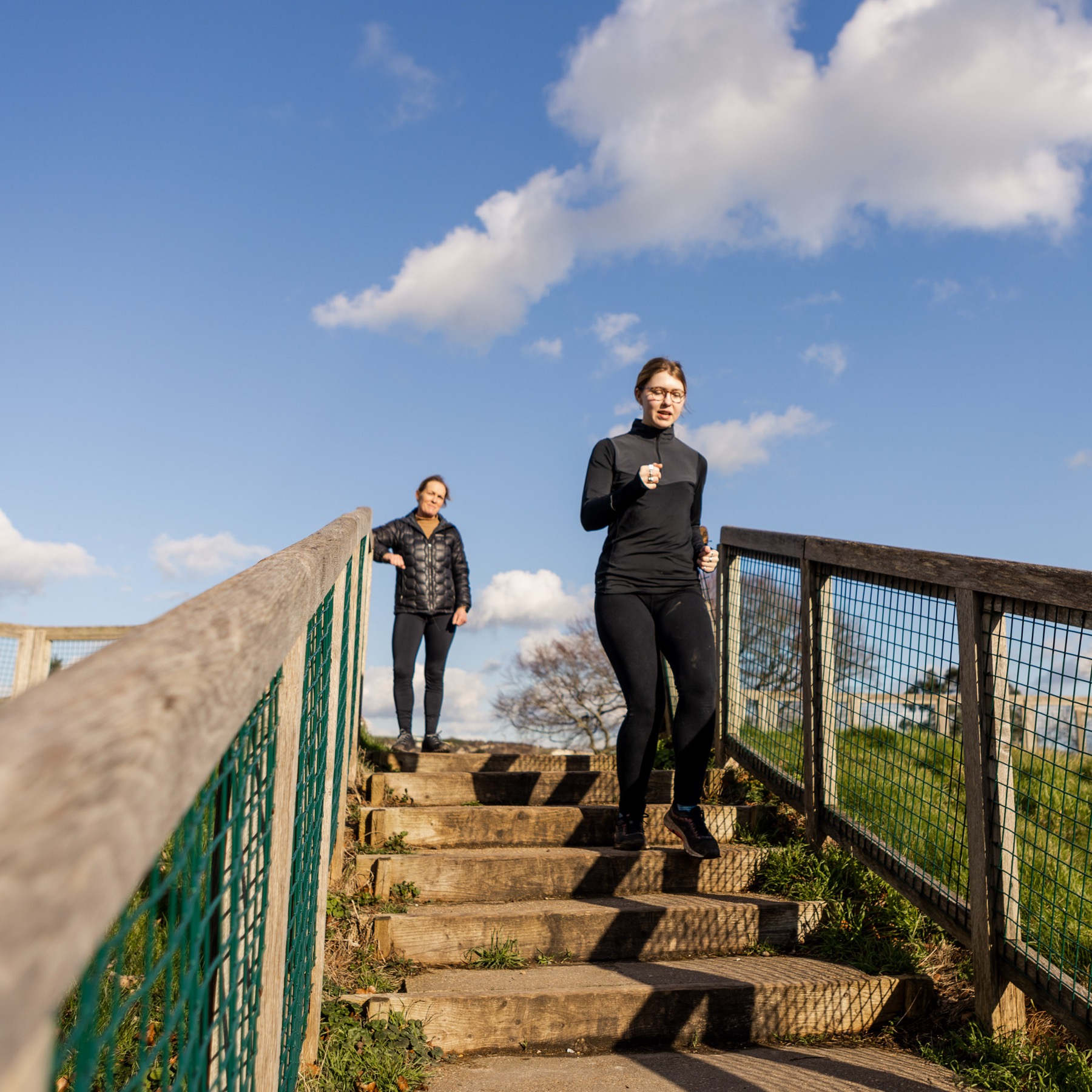 Two women running up a long flight of wooden and stone steps beside a green railing under a blue sky