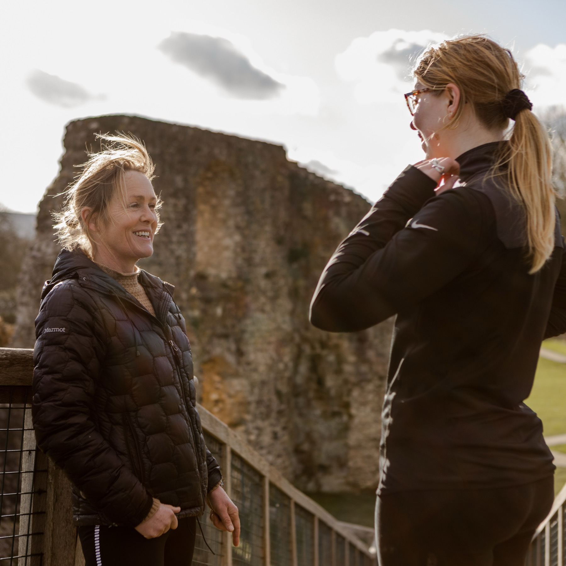 Two women in black training kit talking and laughing in front of the flint ruin of Lewes Castle