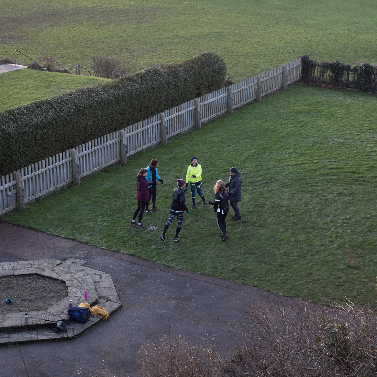A class of about six people on a green enclosed by a hedge and white fence, photographed from above